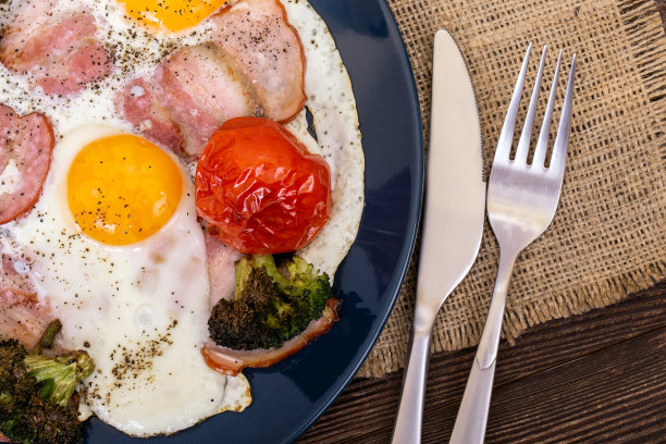 煎早餐用鸡蛋、猪肉、火腿或培根、西红柿和花椰菜盛在盘子里，放在木桌上，用刀叉。宏观照片图片下载