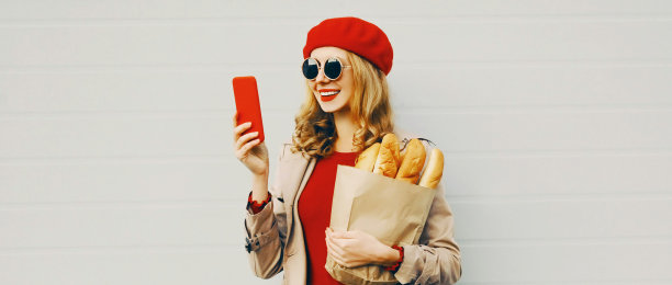 微笑女人的肖像拿着电话和杂货购物纸袋长白面包戴红色贝雷帽在空白的灰色背景图片下载