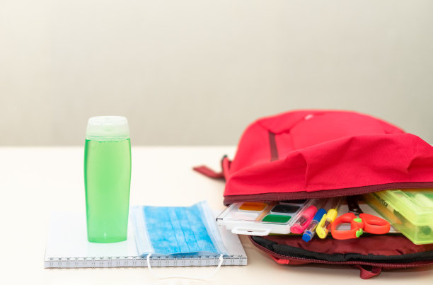 回到学校。教育和社会距离概念。红色背包旁边的桌子上放着医用防护口罩、抗菌洗手液(酒精凝胶)、学习用品。光背景。图片下载