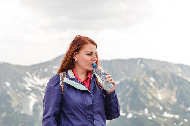 年轻的徒步旅行者在山谷中饮水。山川山水，幸福情感。一个年轻漂亮的红发女人拿着水瓶站在山的背景上。拒绝塑料。图片下载