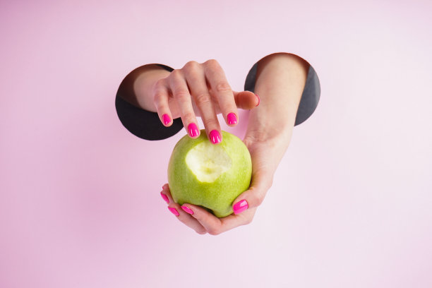 美丽的女人的手拿着一个苹果在粉红色的背景。美丽的美甲概念特写。图片下载