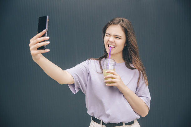 年轻时髦的女人孤立在灰色的蓝色背景。很棒的女孩在自拍，做有趣的面部表情。喝清凉的柠檬水。图片下载