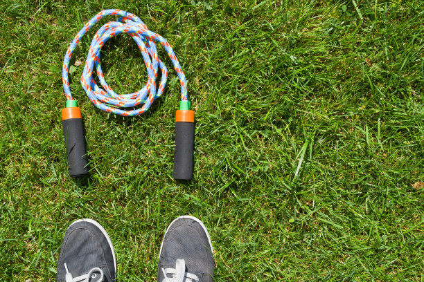 灰色运动鞋和杂色跳绳在明亮的绿色草地背景。在户外跳绳图片下载