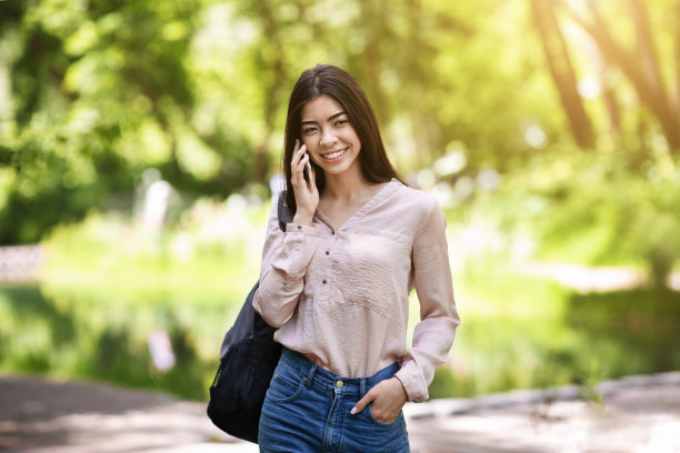 年轻的亚洲女孩背着背包在公园里走路，用手机打电话图片下载
