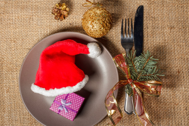 圣诞红帽和礼物装在礼品盒里放在盘子里，刀叉放在餐巾上图片下载