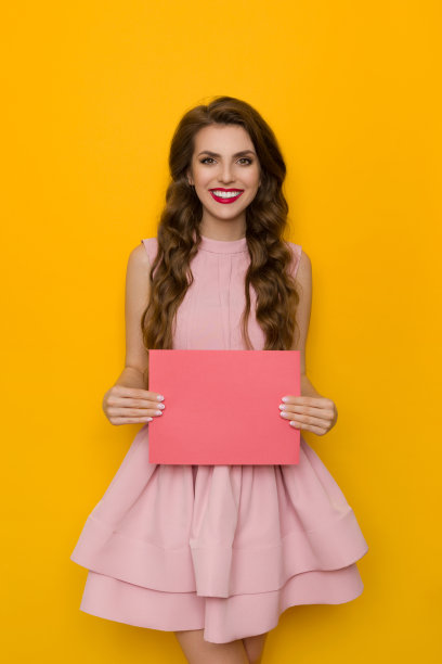 身着优雅的鸡尾酒会裙的可爱微笑的女人拿着一张粉红色的纸图片下载