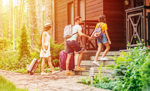 木酒店。这是一家人周末带着行李箱和背包来到树木环绕的木制酒店，父亲牵着女儿的手，带有阳光效果的照片。图片下载