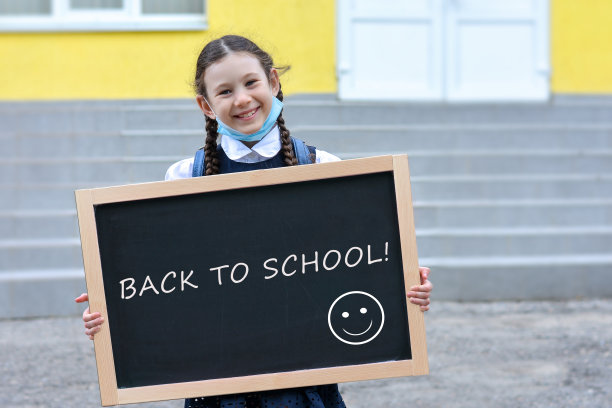 快乐的女孩拿着一块写着“回到学校”的黑板在学校的背景上。学童戴上医用口罩，以保护身体免受流感病毒感染。图片下载