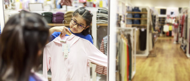 小女孩在购物中心的镜子前试衣服。亚洲女孩微笑快乐的选择和试衣服在镜子前的商店与复制空间横幅。拿着衣服试穿的女孩。图片下载