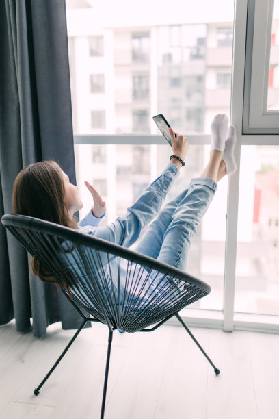 美丽的年轻微笑的女人在她的智能手机自拍坐在一个舒适的大椅子在家里图片下载