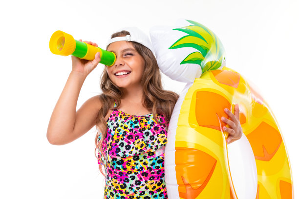 快乐美丽的女孩穿着泳衣，戴着棒球帽，上面有一个充气圈，她通过望远镜在白色的背景上看图片下载