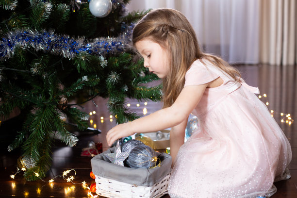 在挂着窗帘的房间里，穿着粉色连衣裙的小女孩用银色和蓝色的圣诞球和珠光宝气装饰圣诞树。寒假的家庭活动。牛年到。照片拍摄。隔离图片下载