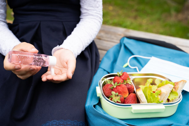 一名身穿蓝色连衣裙的女学生在公园的长椅上，午休时将消毒剂倒在手上。面膜，三明治午餐盒，草莓。蓝色背包上的盒子。冠状病毒感染后的安全措施图片下载