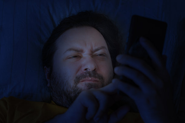 深夜，一位大胡子中年男子躺在枕头上，不高兴地盯着手机屏幕。概念——失眠，阅读社交网络和网上搞笑图片图片下载