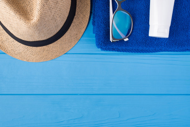 日光浴的概念。顶部上方俯瞰毛巾太阳镜、太阳帽和防晒霜的特写照片孤立在蓝色木制背景与copyspace图片下载