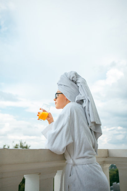 可爱的女孩博主在白色长袍，毛巾和太阳镜在阳台上喝果汁。懒惰的早晨图片下载