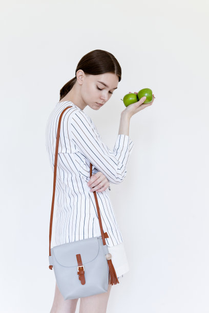 美丽的年轻女子与裸色化妆穿着白色和蓝色条纹的礼服衬衫和白色裙子和浅灰色的袋子拿着酸橙在白色的背景图片下载