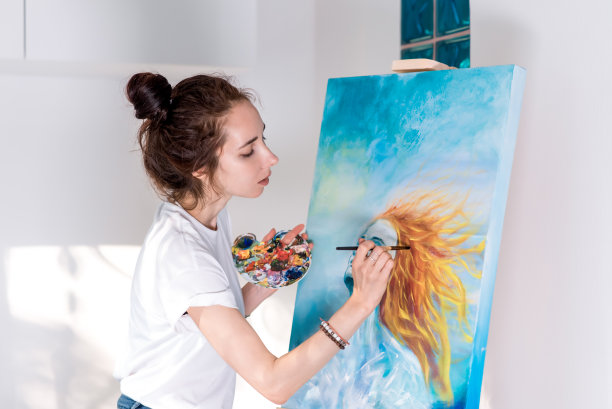 一个女艺术家，画了一个有着红色和橙色长发的女孩在蓝天的映衬下，在她的家里在车间里，她的手拿画笔。背景，手拿画架，调色板和颜料。图片下载