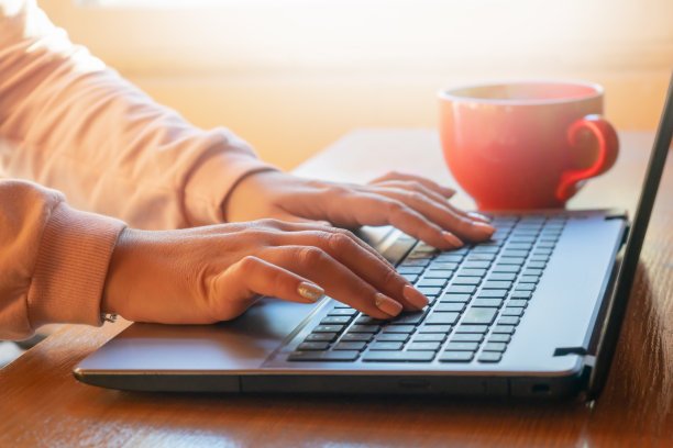 一个女人坐在电脑前工作，旁边有一个红色的杯子。阳光灿烂明亮的光线。图片下载