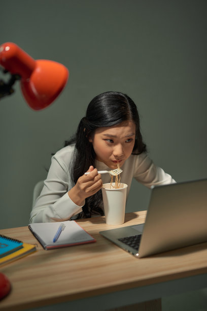 在笔记本电脑前的年轻女性在深夜吃着方便面。图片下载