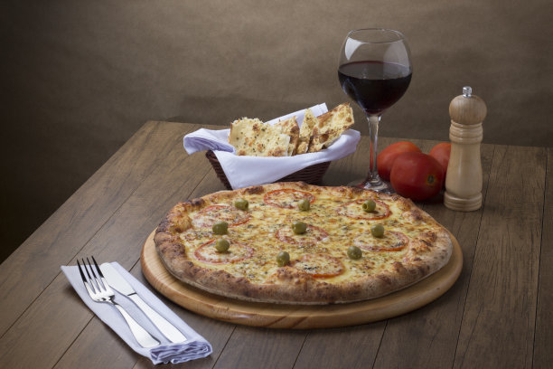 美食照片:马苏里拉奶酪披萨配红番茄和绿橄榄。一杯红酒。叉子和刀放在左边准备品尝。图片下载