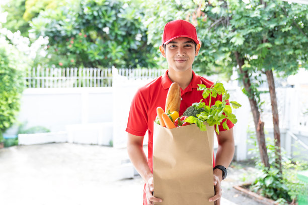 亚洲快递员身穿红色制服，头戴红帽子，手里拿着装着食品、水果、蔬菜的纸袋，微笑着站在家门口。概念快递、外卖图片下载
