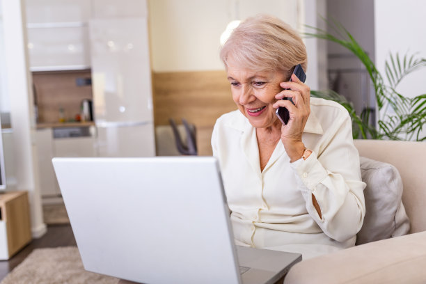 老妇人用笔记本电脑工作，面带微笑，讲电话。使用笔记本电脑的资深女性。老妇人坐在家里，用笔记本电脑和手机聊天，面带微笑。图片下载