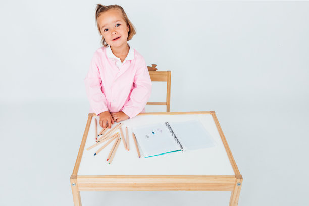 金发女孩，微笑地坐在桌子旁边的椅子上，白色的背景上有笔记本和铅笔。学校的概念图片下载