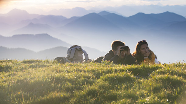 年轻的成年夫妇躺在草地上的山和使用双筒望远镜图片下载