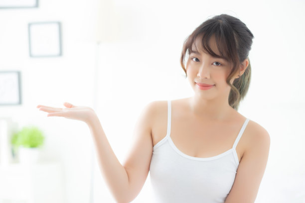 美丽的年轻亚洲女人微笑和展示护肤产品的广告在卧室，美丽的亚洲女孩呈现化妆品的广告与惊喜在手掌在房间。图片下载