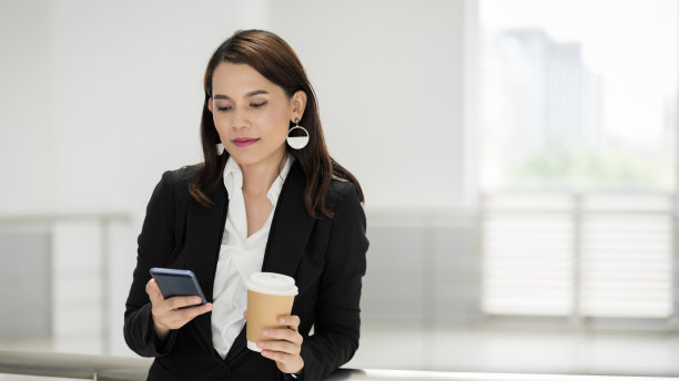 一个自信的女商人的肖像，穿着商务套装，拿着一杯咖啡，在商务大厦的休息时间使用手机。商业库存图片。图片下载