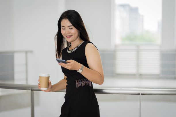 一个自信的女商人的肖像，穿着商务套装，拿着一杯咖啡，在商务大厦的休息时间使用手机。商业库存图片。图片下载