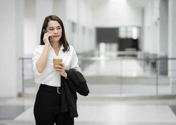 一个年轻的亚洲商业女性在商业大厦里打电话和拿着咖啡的肖像。照片美丽的女孩在休闲套房与电话和一杯咖啡。库存图片图片下载