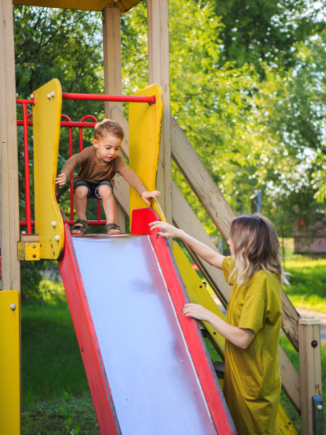 微笑的小男孩和妈妈在公园操场上玩儿童滑梯。关于母亲的夏日照片图片下载