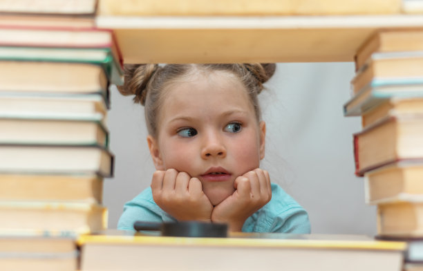 一个惊讶的金发小女生的肖像，她正在翻阅书架上的书。模糊的背景光。回到学校。教育的概念。孩子们的空闲时间。阅读书籍图片下载