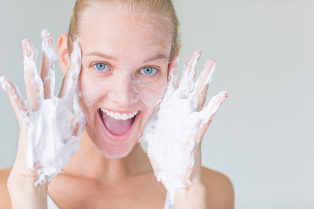 美丽快乐的女人覆盖在肥皂泡沫面部洁面霜和好玩的相机。图片下载