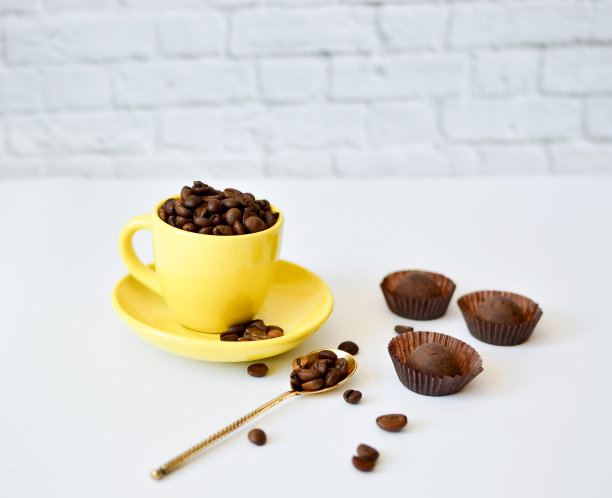 一个黄色的杯子，上面有天然的咖啡豆，白色的背景，还有巧克力。早上咖啡的概念。背景上有花。图片下载