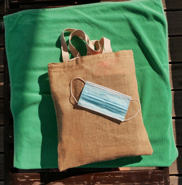 在一个阳光明媚的夏日，浅棕色的沙滩包上放着一个蓝色的医用口罩，浅绿色的棉质沙滩浴巾放在日光浴床上。图片下载
