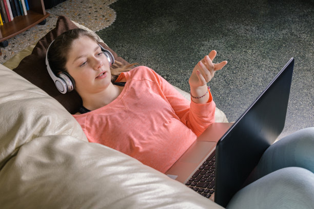 面带微笑的女学生戴着无线耳机，躺在沙发上用笔记本电脑学习或在线交流图片下载