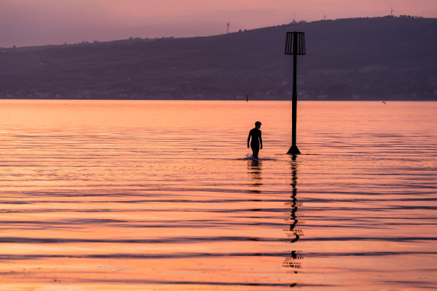 一个穿着潜水服的人的剪影涉水走向低潮标志在平静的水域在日落图片下载