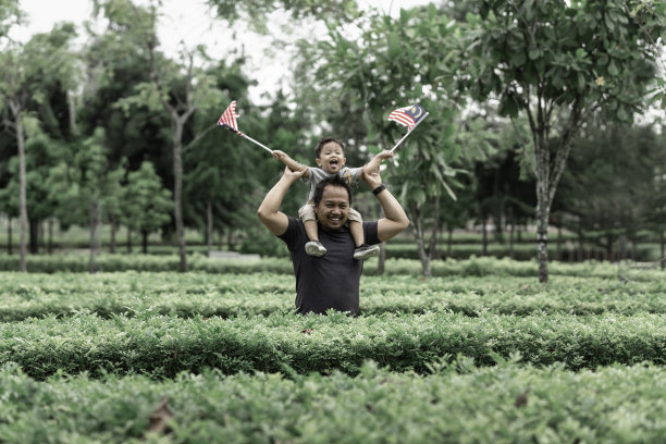 一名马来西亚男孩在公园里举着马来西亚国旗，父亲在他的肩膀上，他在享受早晨的阳光图片下载