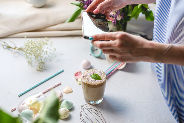 一名妇女正在用手机拍下一杯清晨的达尔戈纳咖啡。白色背景，柔和的颜色和淡紫色的花图片下载