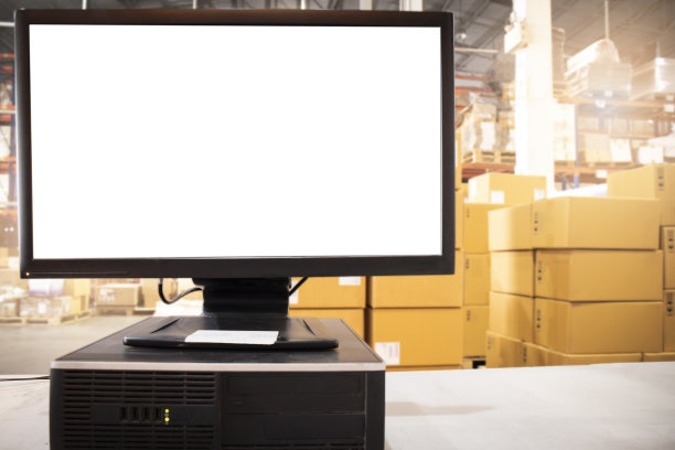 桌面上的电脑空白屏幕和堆放在仓库的纸板箱储存。仓库库存管理。货物出口，货物装运。图片下载