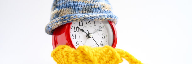 红色圆形闹钟，针织羊毛，蓝色帽子，黄色围巾，白色背景。冬天的时间概念。冬天的季节。舒适和温暖。模拟时间10:10。空间的文本。横幅图片下载