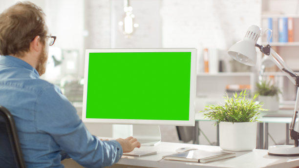 一个年轻的胡子男性使用他的个人桌面电脑与绿色屏幕模拟模板的照片。他在一个明亮的现代办公室工作。图片下载