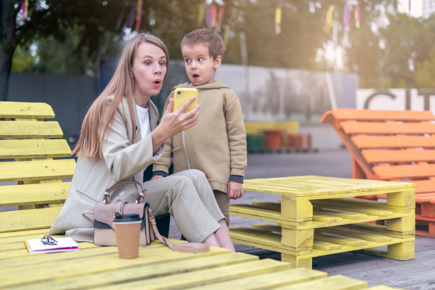 有趣和惊喜的年轻妈妈和学龄前的儿子坐在长凳上使用智能手机，一起玩得很开心。母亲和一个小男孩在城市公园外面享受周末图片下载