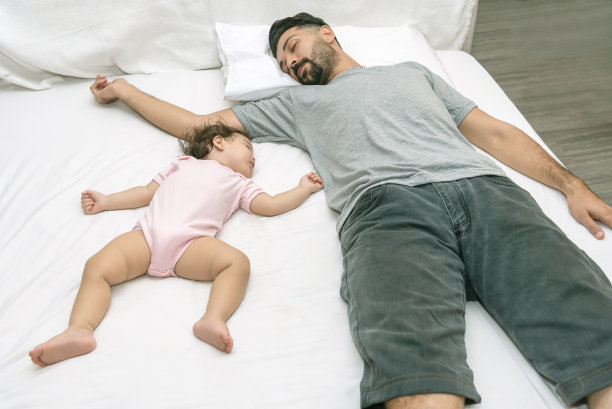 年轻帅气的父亲厌倦了和一岁的小女孩睡觉。大胡子爸爸和宝宝睡在床上。新生儿躺在白色的床上。生育Ccncepts。爱和幸福。图片下载
