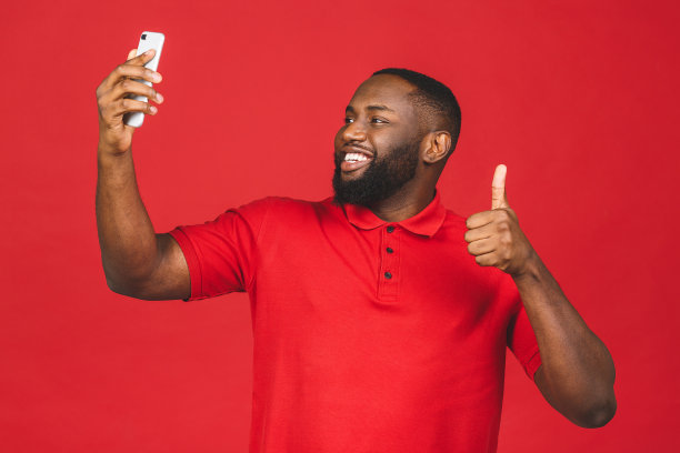 年轻英俊的非裔美国人的肖像站在红色背景下使用智能手机自拍和微笑。图片下载