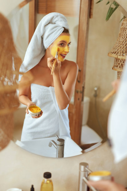 敷面膜的女人。裹着浴巾的模特站在浴室里看着镜子。面部皮肤覆盖美容产品。健康皮肤的日常生活。图片下载