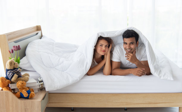 一对有趣的夫妇躺着，说话，躲在卧室的白床上的白毯子里。在家庭关系。早上放松。图片下载